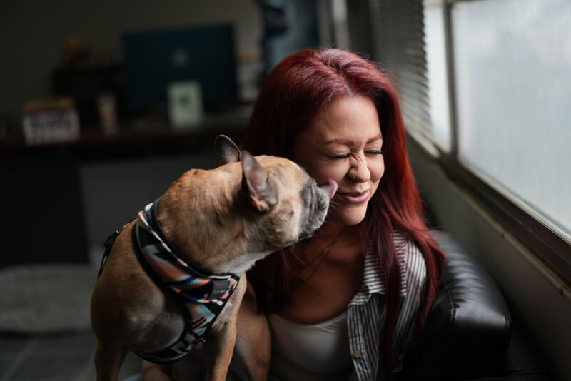 A small dog with a harness licks the cheek of a smiling woman with long red hair sitting by a window, her eyes closed in delight.