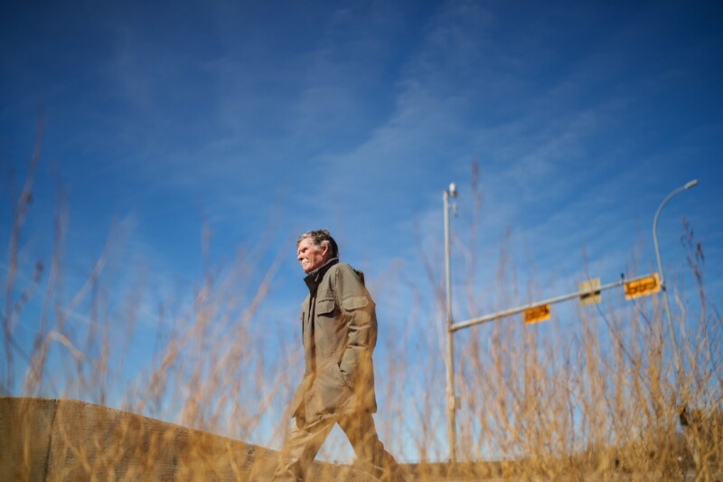 A person walks through a field with dry, tall grass under a clear blue sky. In the background, a streetlight and traffic signals are visible. The individual is wearing a dark coat and appears to be in a thoughtful mood.