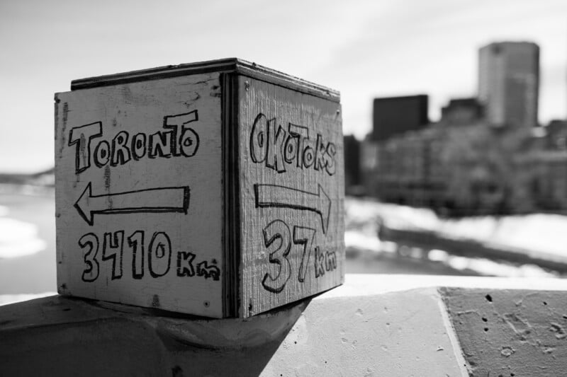 Black and white image of a wooden sign with hand-painted arrows. One points left to “Toronto 3410 km,” the other right to “Ottawa 37 km.” The background features a blurred cityscape under a clear sky.