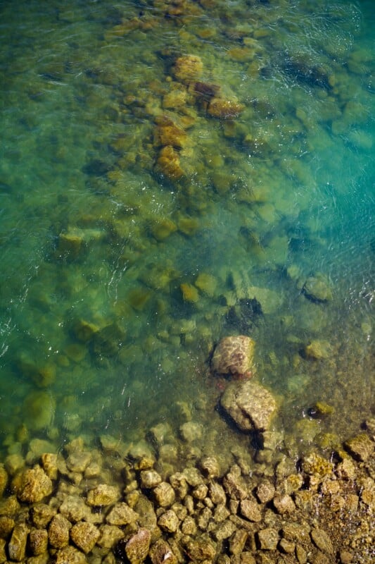 Clear turquoise water with visible rocks of various sizes beneath the surface. The rocks closer to the shore are covered in algae, creating a mix of green and brown hues. The water becomes darker and deeper further from the rocks.