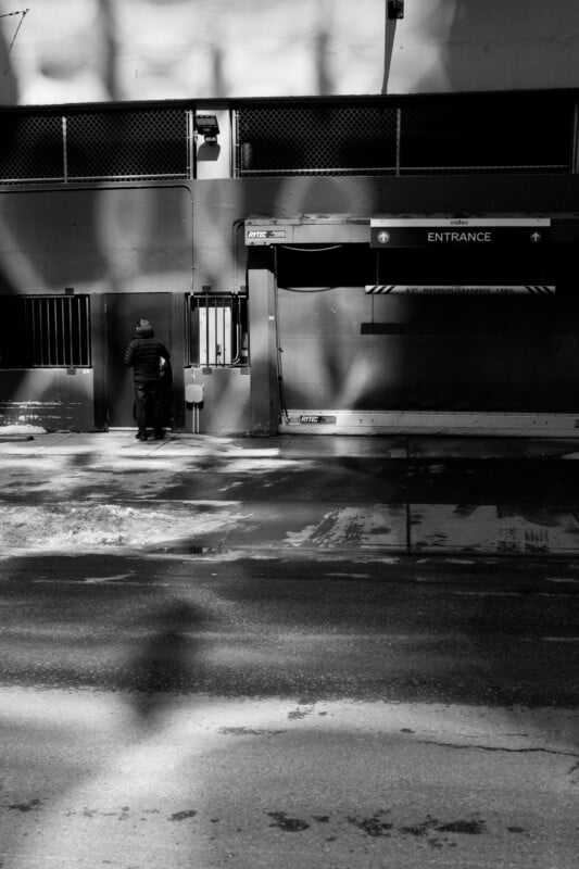 Black and white image of a person standing outside a gated entrance of a parking garage. Shadows of trees create patterns on the ground and building facade. The word "Entrance" is visible above the gate.