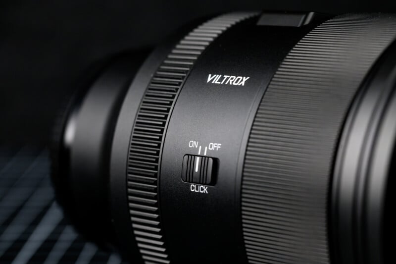 Close-up of a black Viltrox camera lens with detailed focus on the click switch, labeled "ON" and "OFF." The lens features textured grip details on a light grid-patterned background.