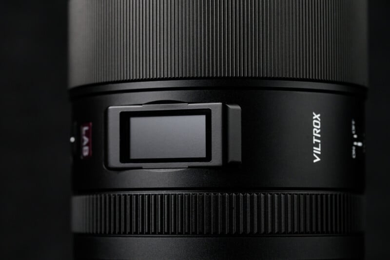 Close-up of a black Viltrox camera lens with textured grip and a square adjustment knob on its side. The lens features sleek, smooth surfaces and a compact design, set against a dark background.