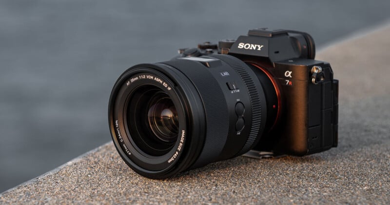 A Sony Alpha 7R mirrorless digital camera with a large lens rests on a textured concrete surface, with a blurred outdoor background.