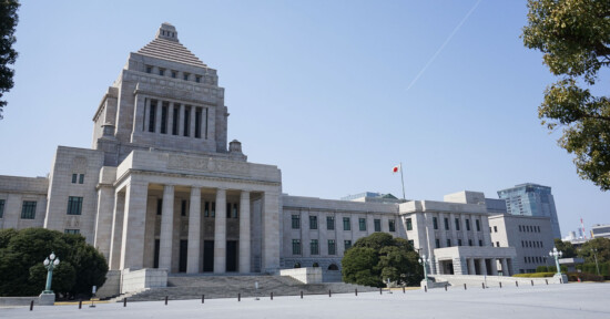 A large stone government building with a central pyramid-shaped tower, columns at the entrance, and a Japanese flag on a flagpole, surrounded by trees under a clear blue sky.