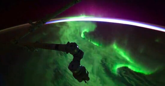 A robotic arm is visible in space with a vibrant aurora glowing green and purple over Earth. The curvature of the planet is outlined against a dark sky, showcasing a stunning space view.