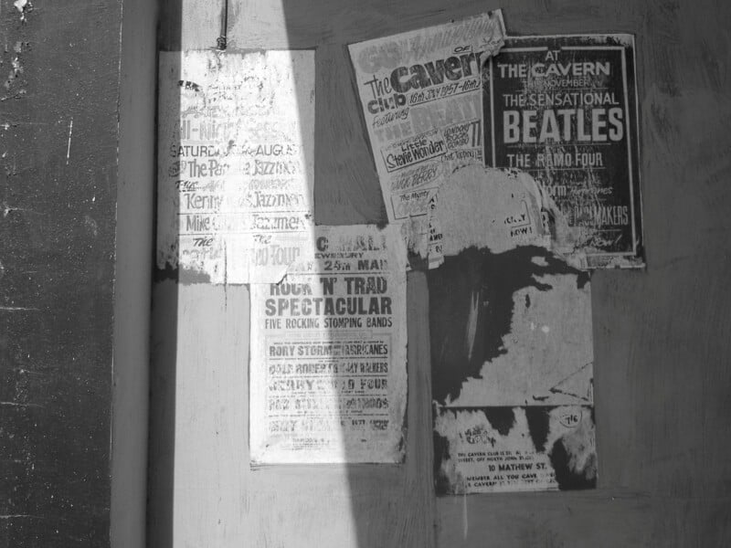 Black and white photo of old, weathered posters on a wall, including one advertising “The Beatles” at The Cavern Club. A diagonal shadow partially covers the posters, adding contrast to the scene.