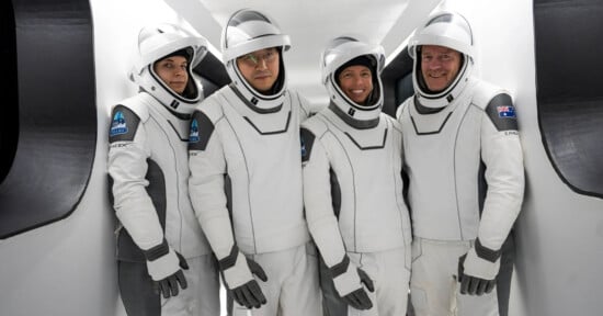 Four astronauts in white and gray SpaceX suits stand inside a spacecraft, smiling and facing the camera. They wear helmets with clear visors and are positioned closely together in a narrow passageway.