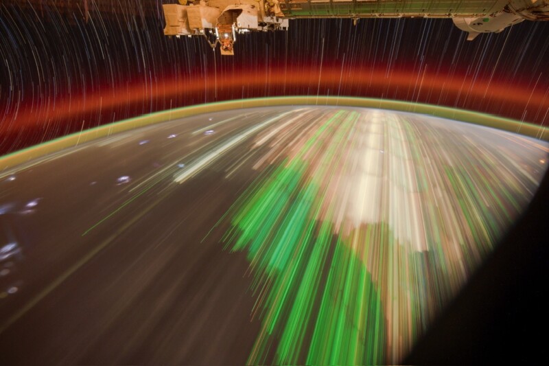 A view from the International Space Station shows colorful light trails over Earth’s atmosphere, with red and green atmospheric bands and star trails appearing in the dark space above the planet.