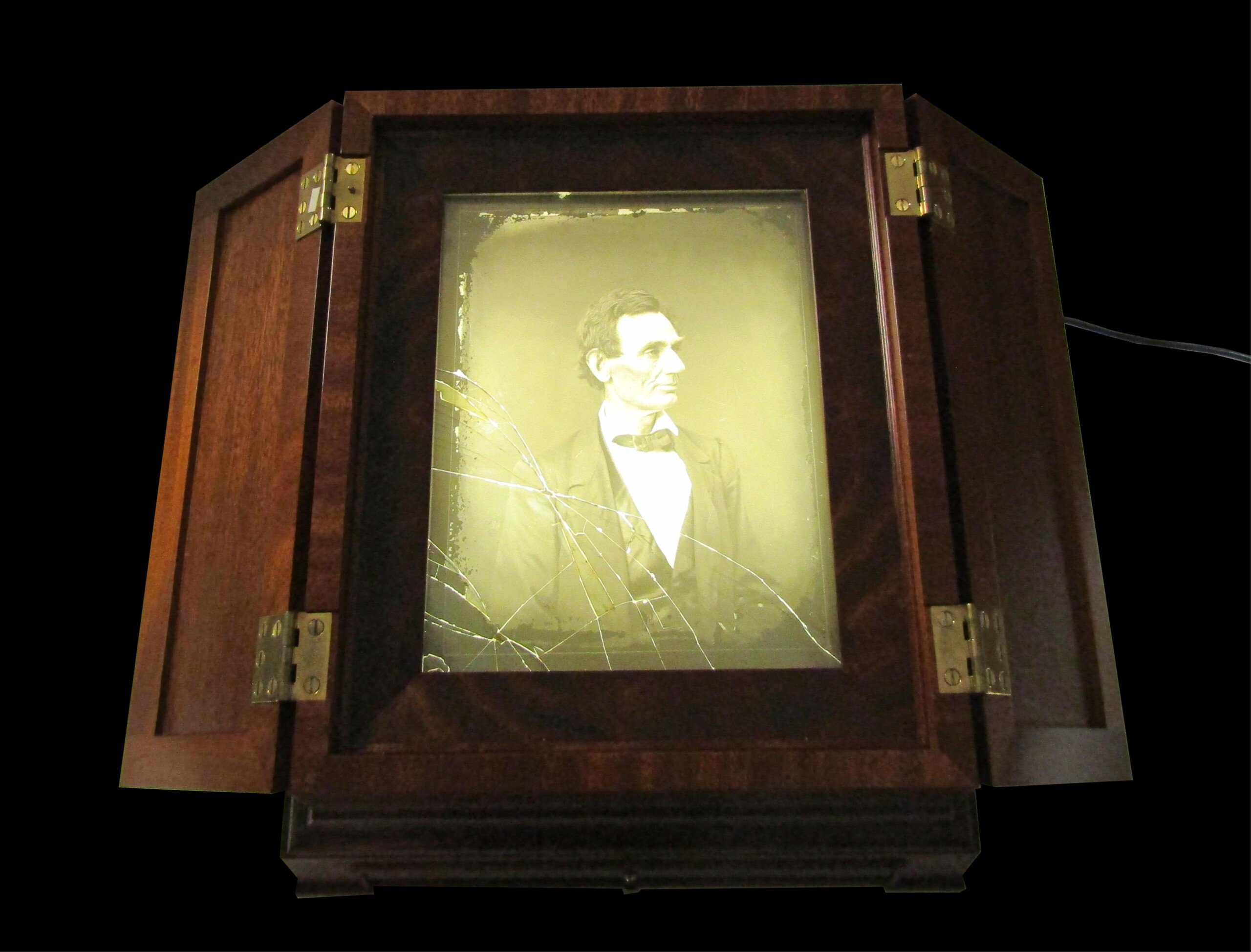 A sepia-toned portrait of Abraham Lincoln is displayed inside a wooden cabinet with open doors and brass hinges. The glass over the portrait is cracked in several places.