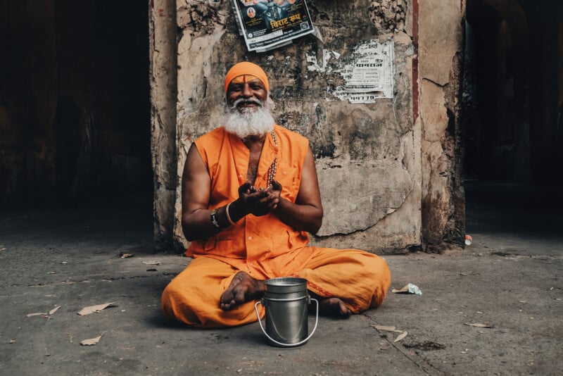 A bearded man dressed in orange robes and a matching headwrap sits cross-legged on the ground, smiling, with a metal container in front of him. The background shows a weathered wall with posters.