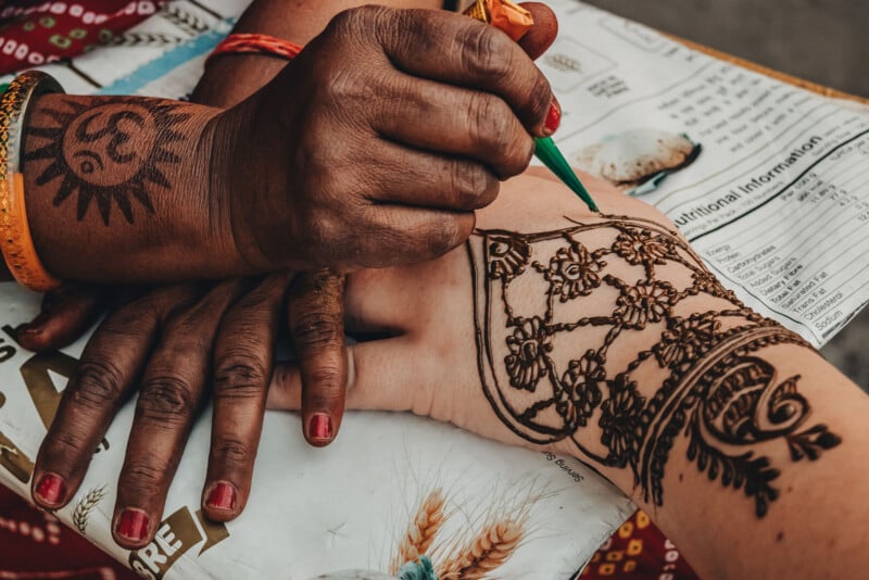 A person is applying intricate henna designs on the back of another person’s hand using a green cone. The artist has a sun tattoo on their wrist and red nail polish. A paper with text is placed below their hands.