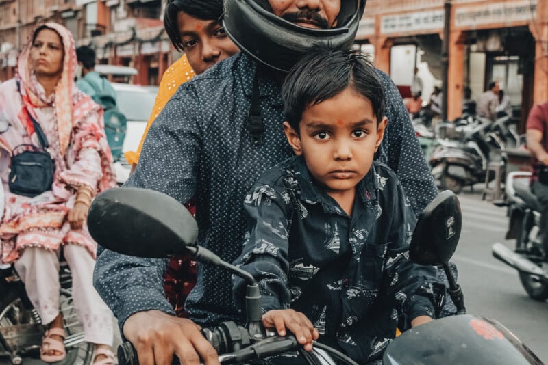 A young boy sits in front of an adult on a motorcycle, looking directly at the camera. The adult wears a helmet. Other people and motorcycles are visible in the background on a busy street.