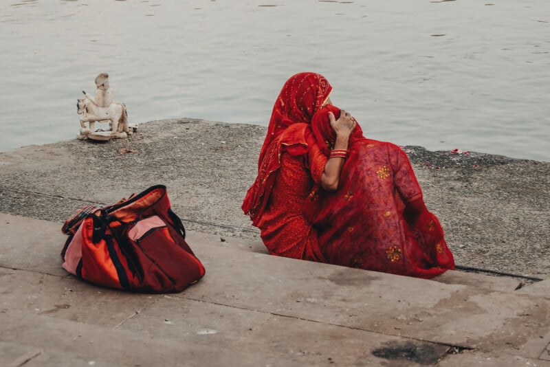 A woman in a red sari sits by a riverside, her back to the camera, holding a child wrapped in a matching red cloth. Nearby are a red bag on the ground and a small white statue near the water's edge.