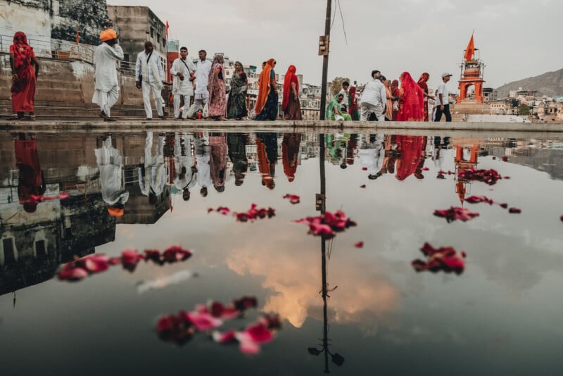 A group of people dressed in colorful traditional attire walk near a temple, reflected in a large puddle with floating flower petals, under a cloudy sky.