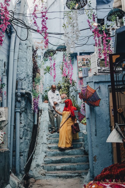 A man and woman stand on blue-painted stone steps in a narrow alley decorated with hanging flowers and colorful umbrellas. The woman wears a yellow sari and red scarf, while the man wears light-colored clothes.