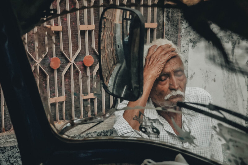 An elderly man with white hair and a mustache sits beside a metal gate, resting his head in his hand with a weary expression. His reflection is visible in a vehicle side mirror in the foreground.