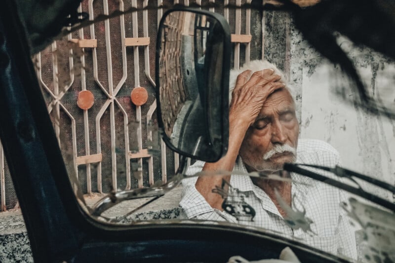 An elderly man with white hair sits behind a metal gate, resting his hand on his forehead with eyes closed. His reflection is visible in a vehicle's side mirror in the foreground.