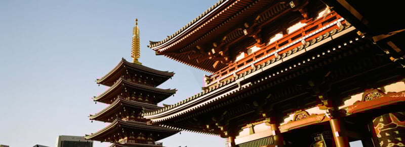 Traditional Japanese temple architecture with a multi-tiered pagoda and a main hall, featuring ornate wooden details and a golden spire against a clear blue sky.