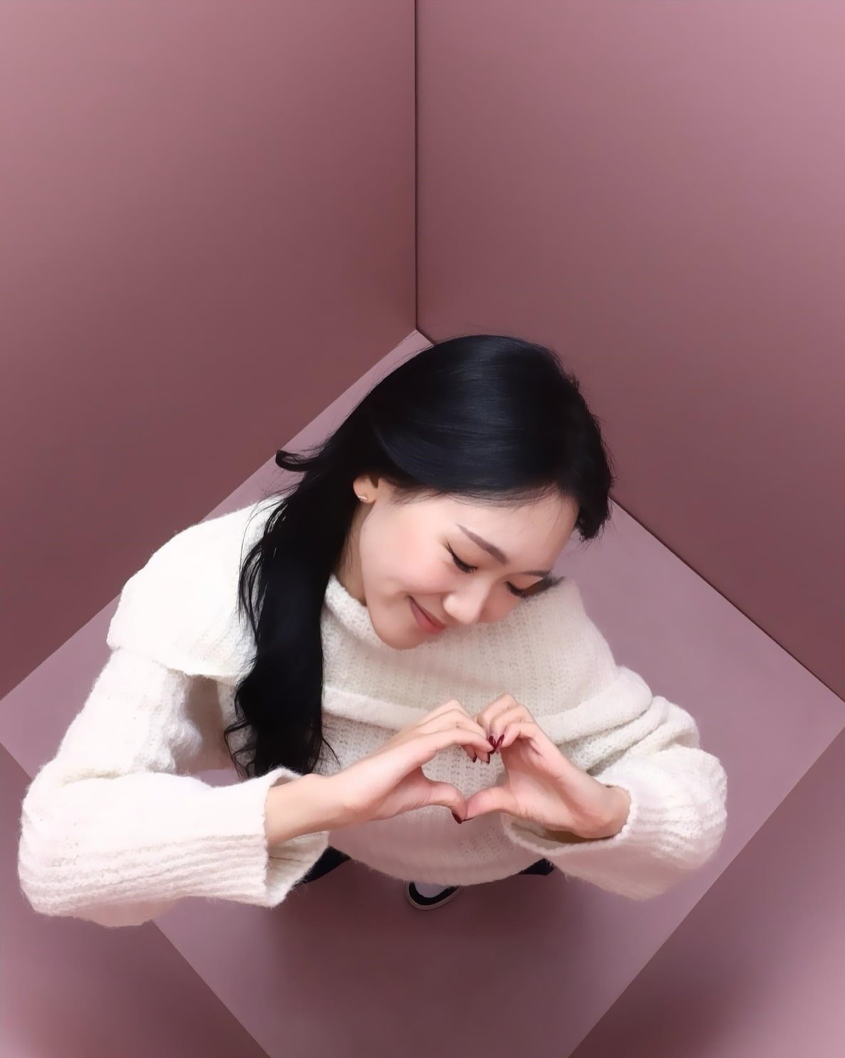 A woman with long black hair wearing a white sweater stands in a pink cornered room, looking down and forming a heart shape with her hands while smiling gently.