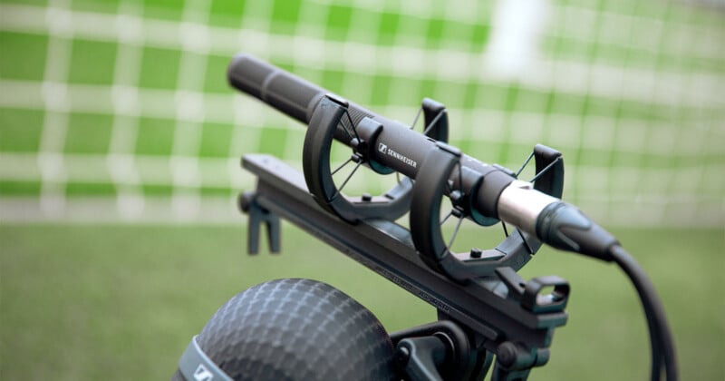 A directional microphone mounted on a stand is set up on a soccer field. The blurred background shows a goal net and green grass, suggesting a sports event or recording session.