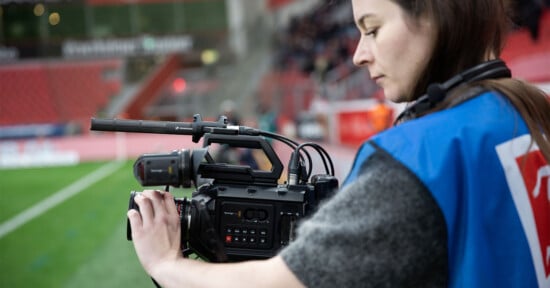 A person operates a video camera at the edge of a sports field. They are wearing a blue vest and have a headset on. The stadium seats and a blurry crowd are visible in the background.