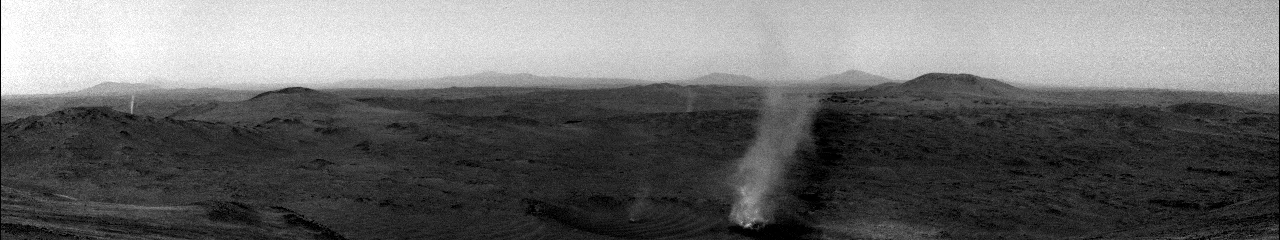 Black and white image of a barren, rocky Martian landscape with scattered hills. A tall dust devil swirls upward near the center, and several distant dust devils are visible on the horizon. The sky is hazy and overcast.