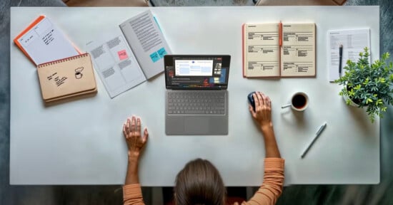 A person sits at a tidy desk with a laptop, notebooks, papers, a pen, a coffee cup, and a small potted plant, working with one hand on the mouse and the other resting on the desk.