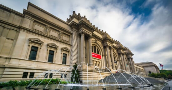 A grand neoclassical building with columns, stone statues, and a wide staircase leading to entrances; water fountains are in front, and a U.S. flag is visible to the right under a partly cloudy sky.