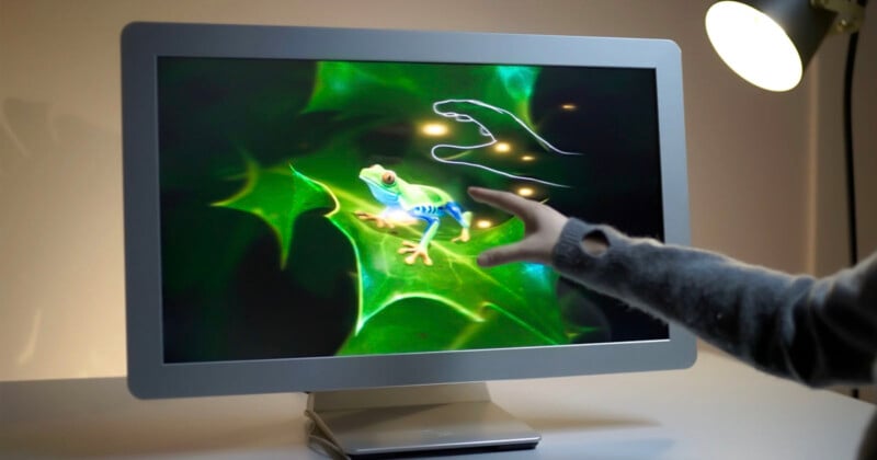 A person touches a computer monitor displaying a vivid image of a frog on green leaves, with a digital hand tracing on the screen. A desk lamp shines in the background.