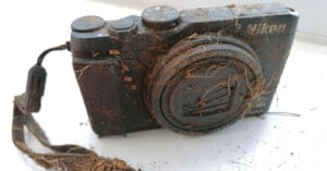 A Nikon digital camera covered in dirt and debris, with mud on the lens cap and body, sits on a white surface. The camera appears weathered and likely found outdoors.