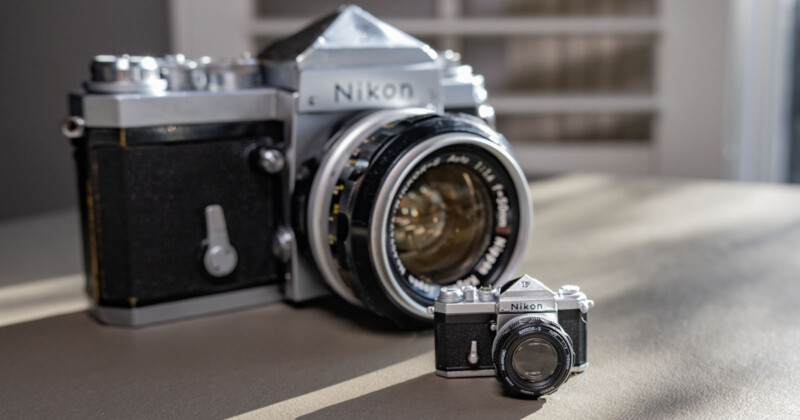 A large Nikon camera with a classic design is placed beside a miniature version on a table. The room in the background is softly lit, highlighting the intricate details of both cameras.