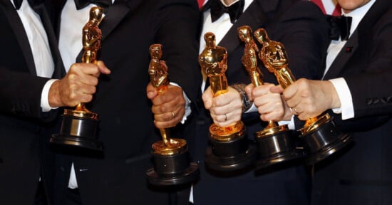 Four people in black suits hold up Oscar statuettes in front of a red and white patterned background, with only their hands and arms visible.