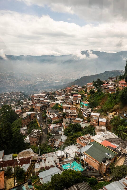 A hillside neighborhood with closely packed houses, many with metal roofs, overlooks a hazy city below. Lush green trees and mountains are in the background under a cloudy sky.