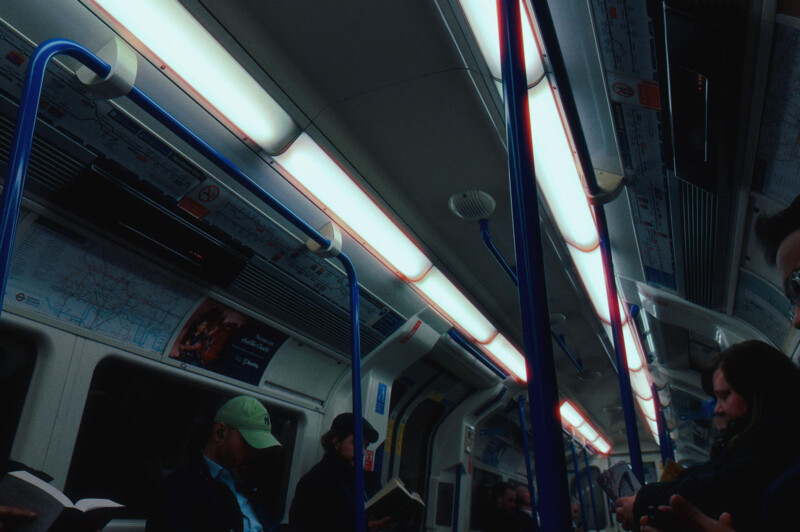 A dimly lit subway car with people sitting and reading. Blue poles and maps are visible on the ceiling. Some passengers wear caps and jackets. The atmosphere is calm and subdued.