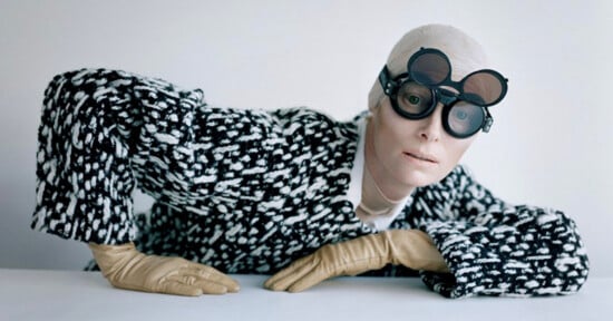 Person in a black and white patterned coat poses against a white background. They wear round, black goggles and tan gloves, with one hand placed on a white surface.