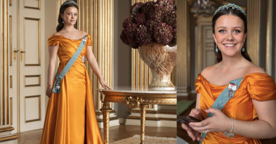A young woman in an orange satin gown with a blue sash and tiara stands in an elegant room with gold accents, a floral arrangement, and holds a phone, smiling.
