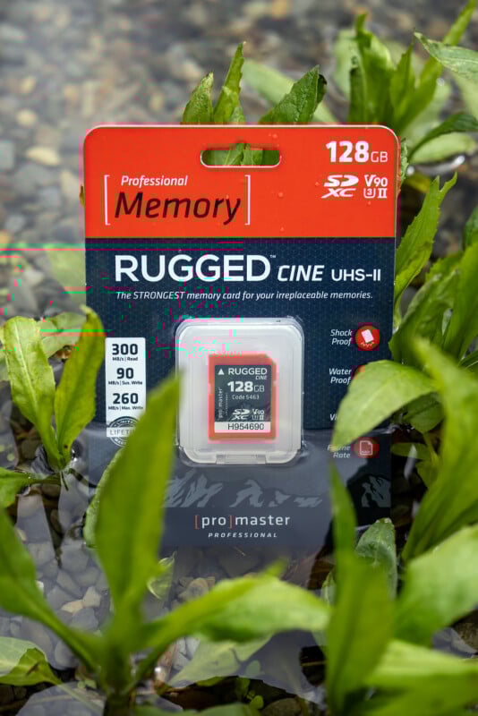 Packaging of a 128GB Rugged Cine UHS-II memory card by promaster, partially submerged in water with green leaves surrounding it. The packaging highlights features like shockproof, waterproof, and fast read/write speeds.