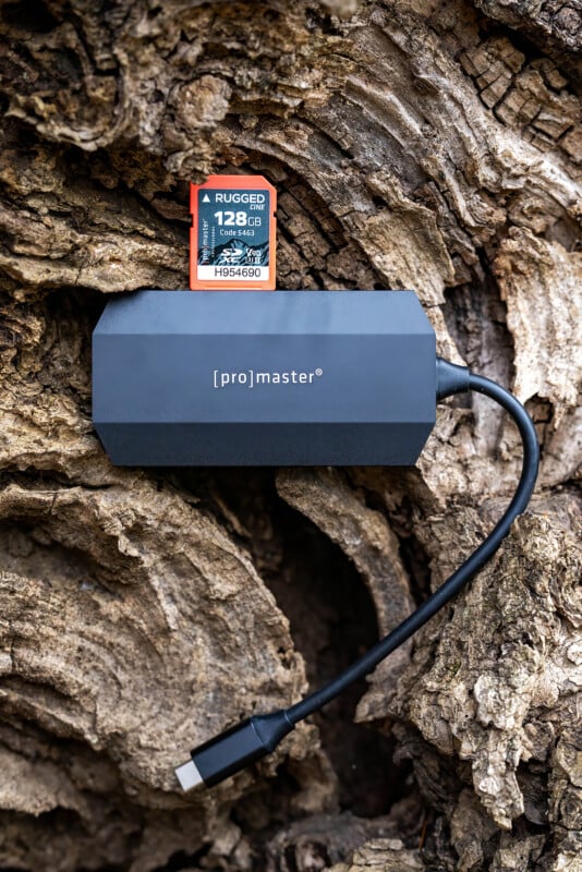 A black ProMaster USB-C card reader with an attached cable lies on textured wood. A 128GB Rugged SD card is placed on top of the reader.