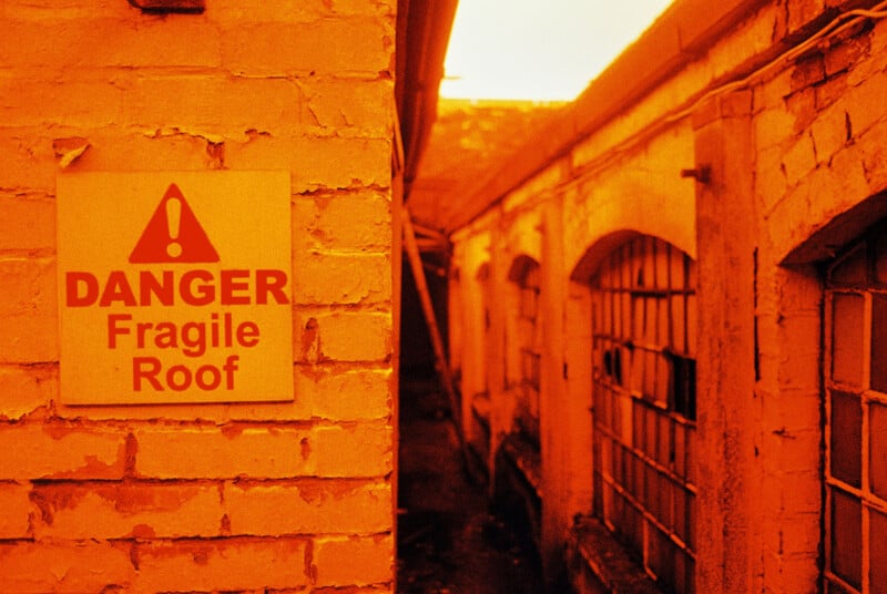 A narrow alley with orange-tinted brick walls. A warning sign on the left wall reads, "Danger Fragile Roof," with a caution symbol. Grid-like windows are visible on the right wall. The scene is illuminated with a warm, orange hue.
