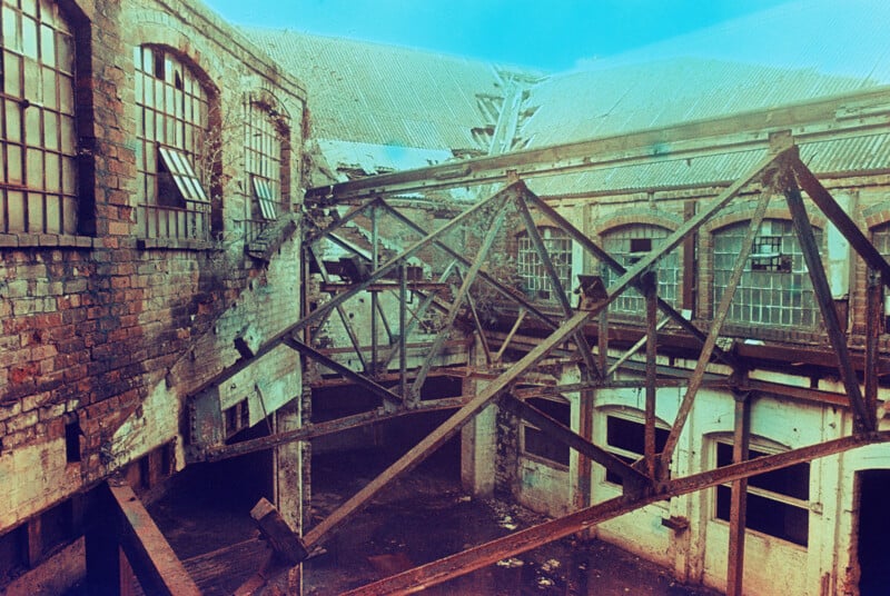 A dilapidated industrial building with broken windows and crumbling brick walls. Exposed metal beams form a crisscross pattern overhead. Debris is scattered across the floor, creating a sense of decay and abandonment in the scene.