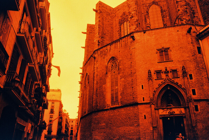 An orange-tinted photo of a narrow street with historical buildings. On the right is a large, ornate stone building with arched windows and a Gothic entrance. On the left, smaller buildings with balconies line the street.