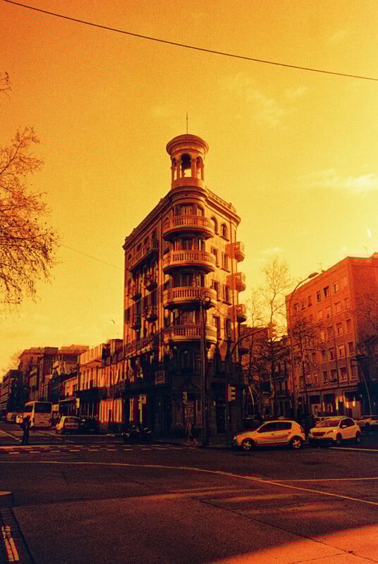 A tall, narrow, vintage building with a tower stands at a street corner under a vivid orange sky. Cars are parked nearby, and bare trees line the street. The image has a warm, sepia-like tone.