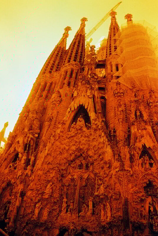 A dramatic, orange-tinted image of the Sagrada Família's intricate stone facade, showcasing soaring spires and detailed carvings under a hazy, yellowish sky. Scaffolding is visible on part of the structure.