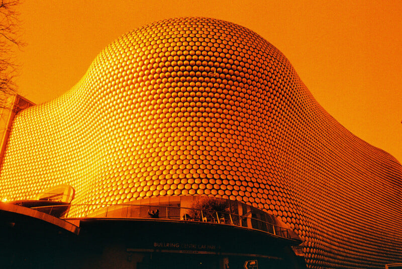 A striking modern building with a wavy structure covered in circular metal discs, set against an orange sky. Its unique, futuristic design features rounded, flowing shapes and reflective surfaces.