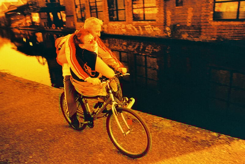 Two people joyfully ride a bicycle along a riverside path during golden hour. The person in front pedals, while the other hugs them from behind. The scene is warm with an orange hue, and old brick buildings line the opposite side of the river.