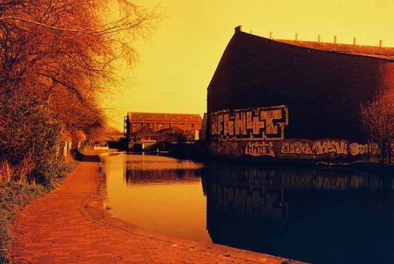 A canal scene with a brick path running alongside the water. A large, industrial building with graffiti is on the right, reflected in the canal. The sky and surroundings have an orange tint, giving a vintage, surreal effect.