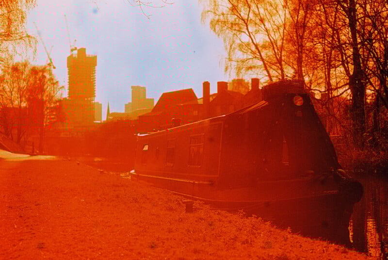 A narrowboat is docked along a canal under red-tinted light. Leafless trees line the path, and tall buildings are visible in the background. The scene has an ethereal, dreamy atmosphere.