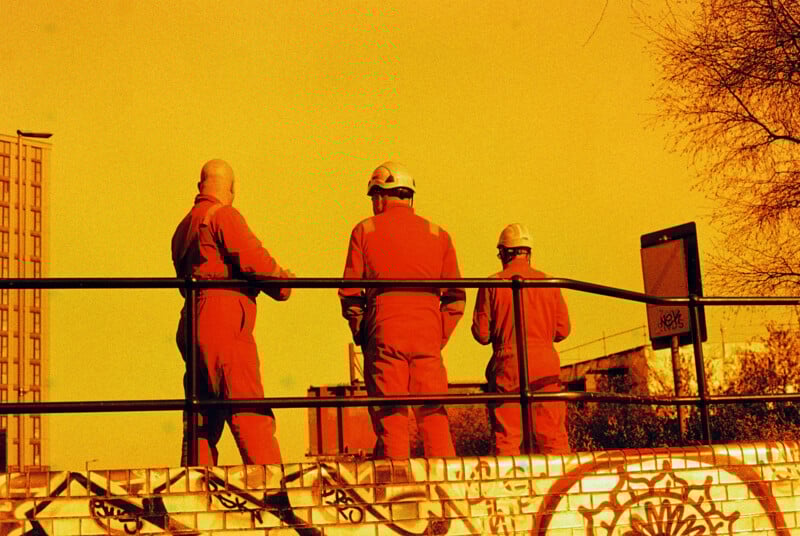 Three construction workers in orange suits and hard hats stand on a graffiti-covered structure. The sky is a vivid orange, casting an unusual hue over the scene. A building is partially visible in the background on the left.