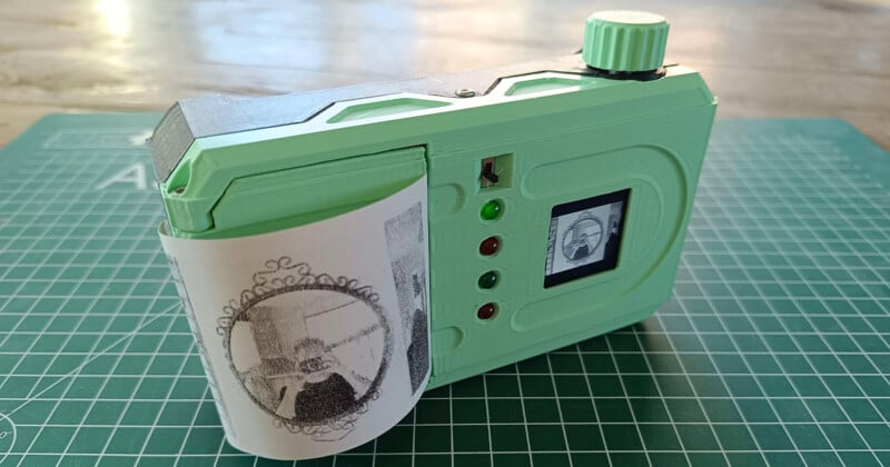 A green instant camera with a retro design sits on a cutting mat. It has a printout showing a portrait emerging from its side. The camera has a few red and green buttons, with a small display screen on its body.